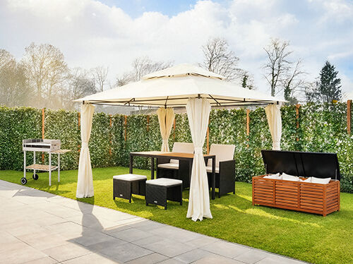 Outdoor garden setup featuring a cream gazebo with curtains, a dining table and chairs, stools, a barbecue trolley, and a wooden storage box on a green lawn. Click to see the products in the collection.